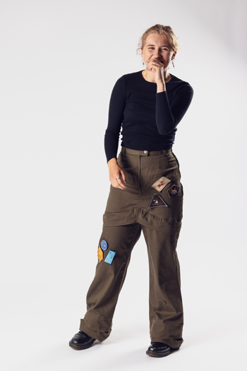 Person wearing a black long-sleeved t-shirt and olive-green cargo pants with embroidered patches, smiling with a hand over their mouth.