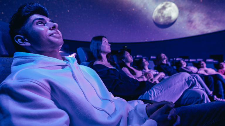 Adults sit on reclining chairs in the Melbourne Planetarium at Scienceworks with projections on the Planetarium dome of the Moon and the Milky Way galaxy