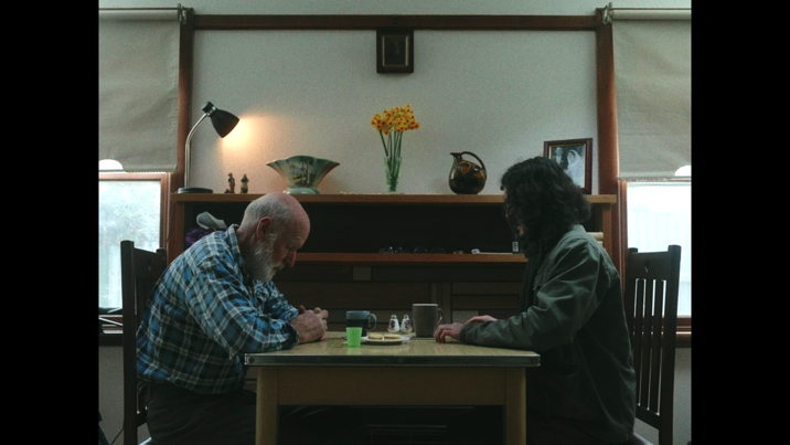 An elderly man and a younger person sit across from each other at a wooden table in a dimly lit room, heads bowed. A vase of flowers can be seen on shelves behind them.