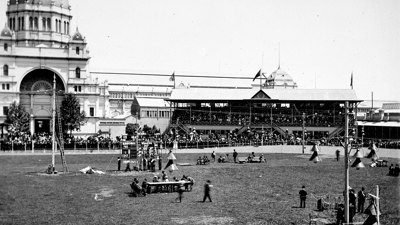 A view of the crowd, arena, officials and grandstand at the bicycle sports day held on ANA Day. The Exhibition Building can be seen in the background.