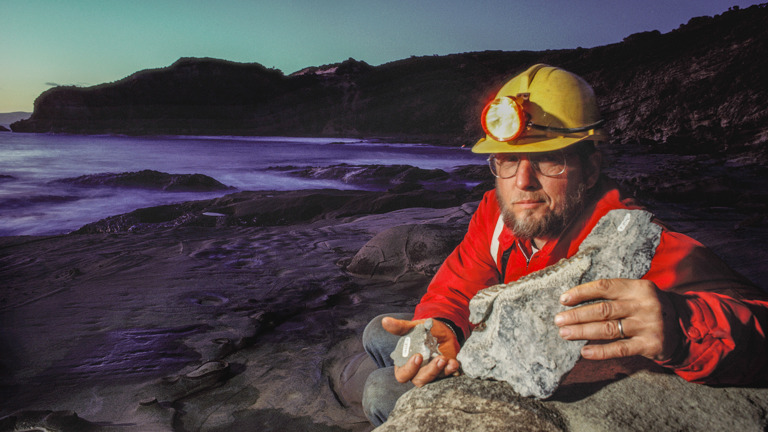 A man crouching down on a beach, wearing a hard hat, holding two rocky fossils