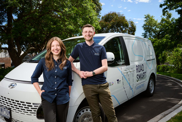 Two people dressed in a Road Smart Interactive uniform leaning up against a van, smiling directly at the camera
