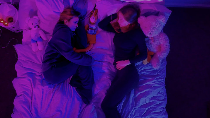 Film still showing two teenaged girls in dark clothing, lying on a bed in purple and blue lighting, surrounded by stuffed animals and pillows.