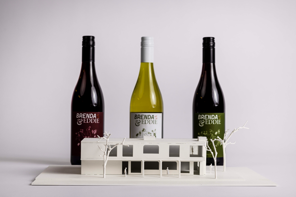 Three wine bottles with different coloured labels behind a white architectural model of a two-storey building with trees and small human figures.