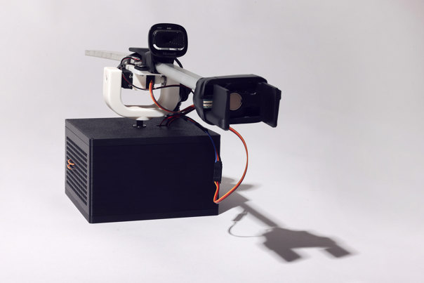 Robotic device with a black, rectangular base unit, white articulated arm mechanism with exposed wiring, top-mounted camera lens and a mobile-phone holder.