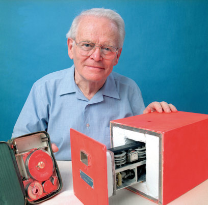 A man wearing glasses and a blue shirt looking at the camera, he is holding a red box and case containing cogs.