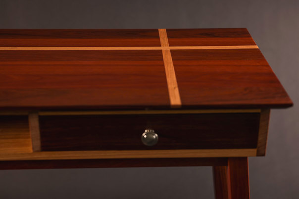 Close-up view of a wooden table that features a cross design on its top and an inset drawer with a silver handle.