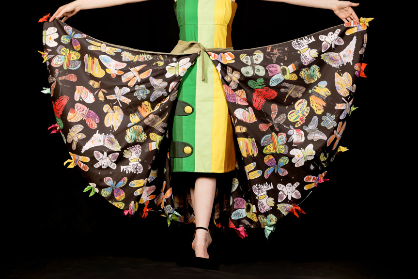 Person wears a cape adorned with butterfly illustrations. A green, yellow and brown shift dress is worn underneath.