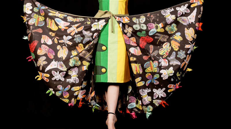 Person wears a cape adorned with butterfly illustrations. A green, yellow and brown shift dress is worn underneath.