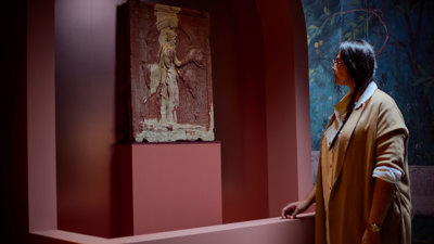 Visitor standing beside a carved stone relief set within a curved, terracotta-colored exhibition display in ROME: Empire, Power, People exhibition in Melbourne Museum.. 