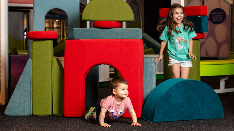 Two Children playing with cushions they have used to make a cubby house in tinkertown.