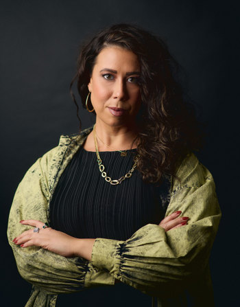 Kimberley stands with the hands crossed. Kimberley wears a black top and washed-green denim jacket. 