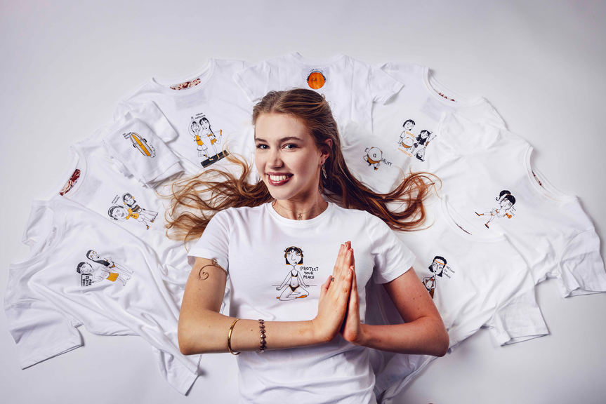 Smiling person lying on a white background with arms in a prayer position, surrounded by white T-shirts with hand-drawn illustrations.