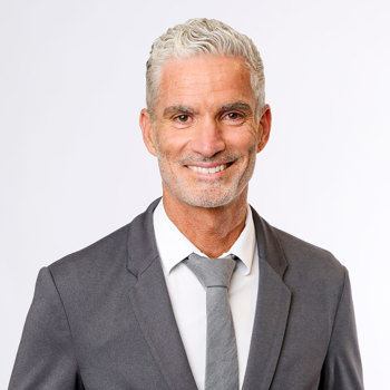 On a white background, Craig Foster smiles at the camera. Craig wears a grey suit with white shirt and light grey tie. 