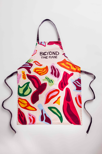 Apron with a colourful abstract pattern and black neck and waist ties, featuring the text "BEYOND THE PLATE" in bold black letters.