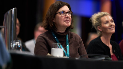 Seated woman with mid length dark brown hair and glasses, looking at someone not in the photograph. Blonde haired woman in the background.