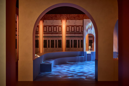Arched gallery space inspired by a Roman bathhouse, with columns, projected light patterns, and a small sculpture in an alcove in ROME: Empire, Power, People exhibition in Melbourne Museum.. 