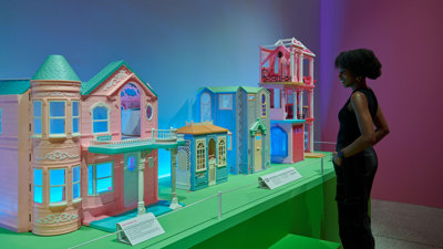 Museum visitor viewing a display of pastel-coloured dollhouses illuminated by vibrant exhibition lighting.