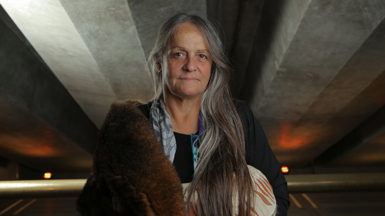Portrait of a woman in a carpark wrapped in a possum skin cloak.