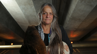 Portrait of a woman in a carpark wrapped in a possum skin cloak. 