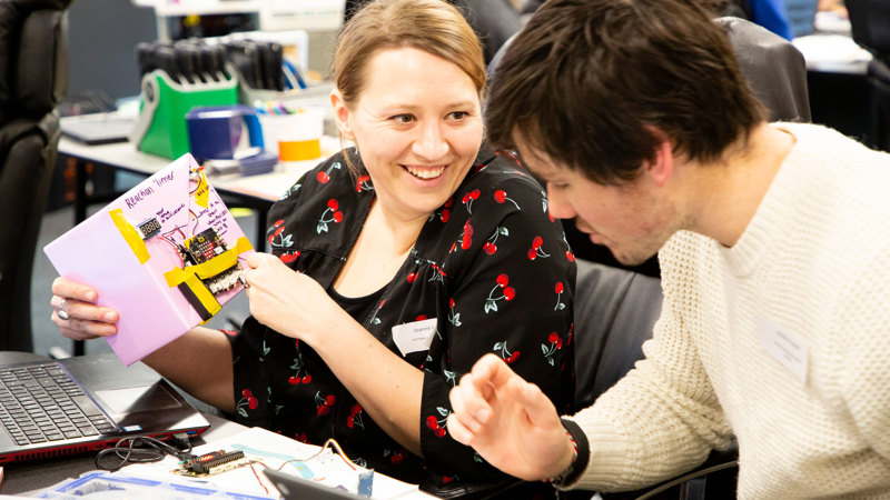 Two people sitting at a desk collaborating. There is a computer and other electronic components visible.