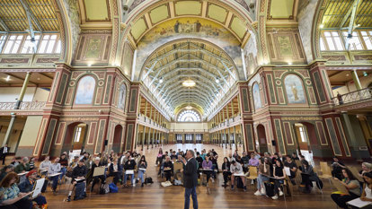 A group of budding artists sis on chairs under the dome of the Royal Exhibition Building for a drawing class.