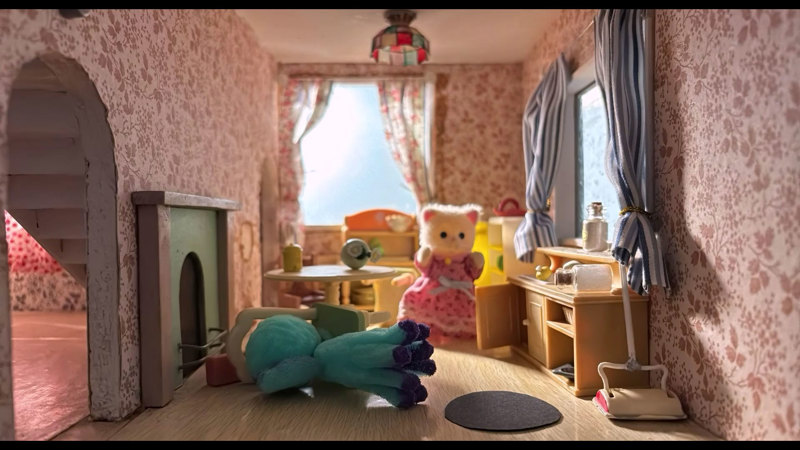 Stop-motion animation scene showing a miniature bedroom with floral wallpaper. A white cat figure wearing a pink dress can be seen amid furniture in disarray.