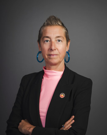 On a dark grey background, Jana looks straight into the camera standing with their hands crossed. Jana wears a black blazer with an Indigenous flag pin, pink turtleneck and blue hoop earrings.   