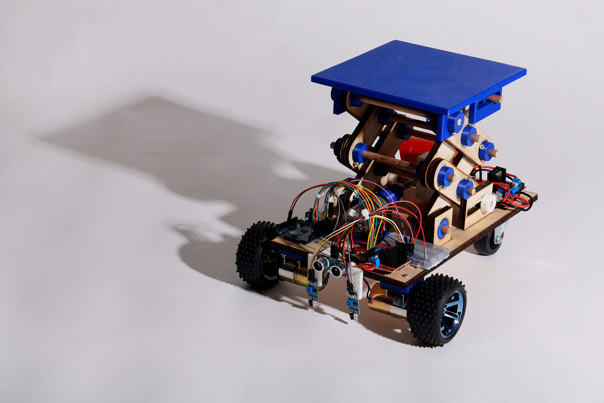 Robotic vehicle with blue platform and wooden scissor-lift mechanism on a wooden base, featuring visible circuit boards, coloured wiring, and black wheels.