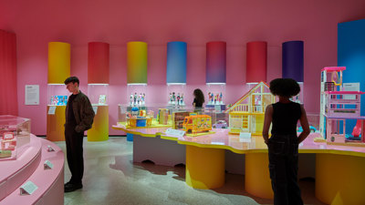 Three museum visitors in a colourful gallery room, looking at various Barbie exhibits 