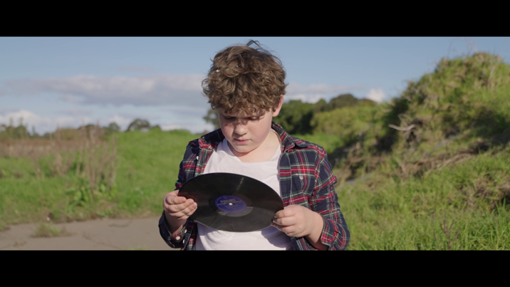 Young boy in a flannelette shirt stands examining a vinyl record, against a backdrop of green hills and blue sky.