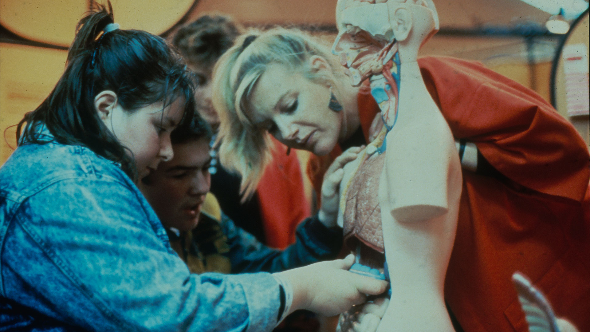 A teacher speaks to a group of children crouching in a circle around a small wooden chest.
