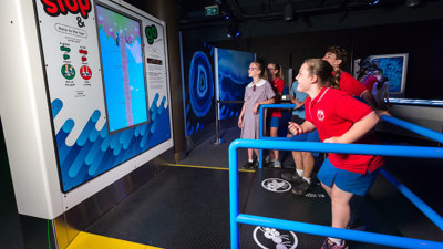 A group of school‑aged children participates in an interactive exhibit featuring a large vertical screen with “stop & go” instructions. Some children stand on a platform marked with graphics, while one child appears ready to run or move as part of the activity. Blue railings guide participants along the path, and the exhibit displays colourful graphics resembling water or waves. 