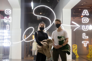 Two people wearing masks stand in front of a neon sign with ‘CHILD’ and ‘ADULT’ illuminated on either side of them. 