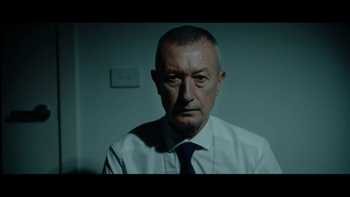 Film still showing an older man in a white shirt and dark tie with an intense expression, lit in cool tones with a door visible in the background.