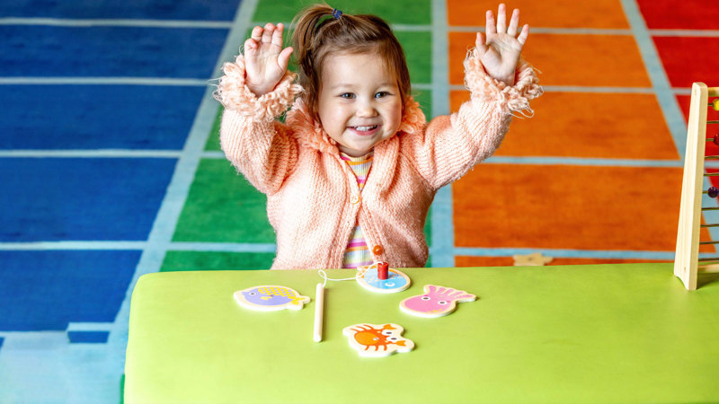 Little girl on a colourful rug smiling with her arms in the air. She is playing with a magnetic fishing rod and wooden sea creatures. 