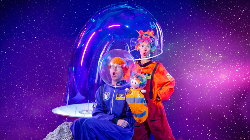 Two performers in bright orange and blue space suits with bubble helmets sit on an asteroid as it zooms through space.