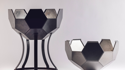 Geodesic metal sphere with hexagonal panels in dark tones, split horizontally in two. The bottom half rests on a curved black-metal stand while the top half sits alongside. 
