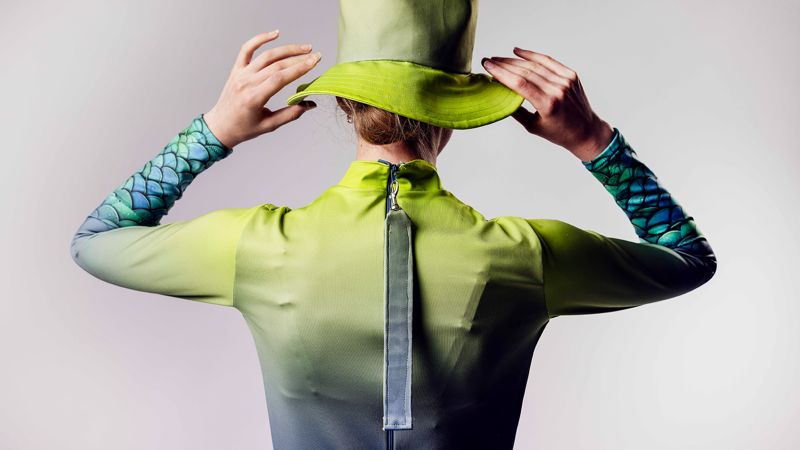 Person seen from behind wears a green long-sleeved rash top with fish-scale patterned sleeves and a matching bucket hat. 
