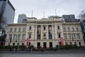Melbourne’s Immigration Museum, a classical building with a sand-coloured exterior, red flags flying in the foreground and two banners with a man and a woman’s face draped beside the entrance.