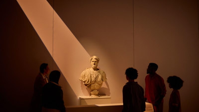 Group of visitors standing around a marble Roman bust on a plinth under dramatic overhead lighting in ROME: Empire, Power, People exhibition in Melbourne Museum.. 