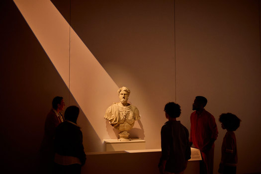 Group of visitors standing around a marble Roman bust on a plinth under dramatic overhead lighting in ROME: Empire, Power, People exhibition in Melbourne Museum.. 