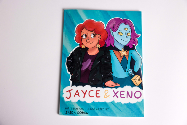 Illustrated book cover featuring two characters: one has red curly hair, a black jacket, and star earrings. The other has blue skin, three eyes, and a cape.