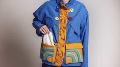 Model wearing a blue jacket with colourful oversized buttons and an orange pleated placket, featuring pockets with decorative embroidery.