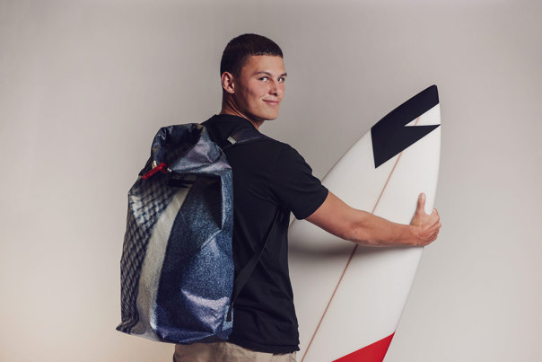 Model wearing a large black backpack with printed graphic design, holding a white surfboard with black and red angular details.