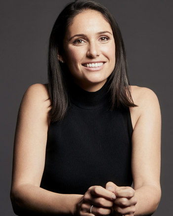 On a dark grey background, Kate Gill smiles at the camera. Kate wears a black sleeveless turtleneck. 