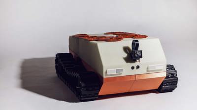 3D-printed tracked vehicle in cream and terracotta colours featuring a sloped top with solar panels, dual tank treads, and visible electronic components on the front.