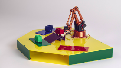 Small robotic arm on a yellow hexagonal platform, sorting model trucks in a variety of colours.