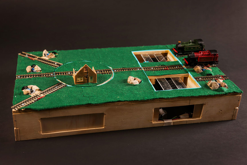 Model trains, train tracks and a small house sit atop a wooden box with a green felt top. System components are visible inside the box.