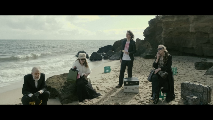 Four elegantly dressed people sit and stand on a sandy beach, surrounded by luggage and crates. Waves and a rocky outcrop are seen in the background.